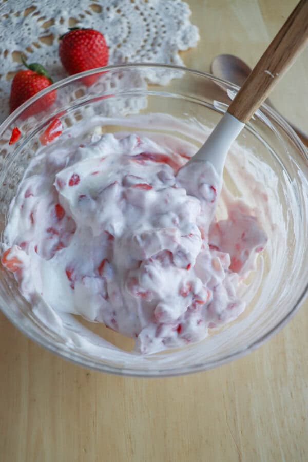 A glass bowl filled with a creamy strawberry mixture, showcasing chopped strawberries mixed with yogurt. A wooden-handled spoon rests inside the bowl. Two whole strawberries sit on a lace doily in the background.