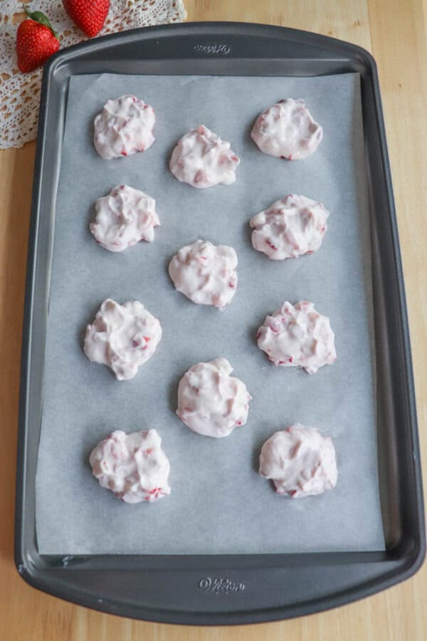 A baking tray lined with parchment paper holds twelve dollops of strawberry yogurt mixture. Two fresh strawberries and a lace doily can be seen in the background on a wooden surface.