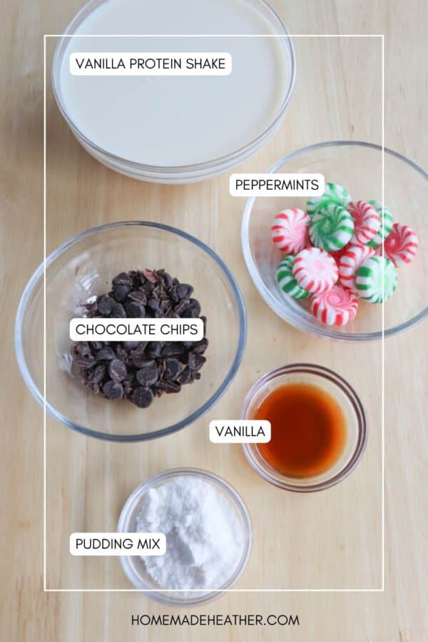 An overhead view of ingredients on a wooden surface: a bowl of vanilla protein shake, a bowl of peppermints, a bowl of chocolate chips, a small bowl of vanilla extract, and a bowl of pudding mix. Labels identify each ingredient.