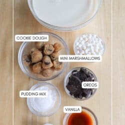 Ingredients for a vanilla protein shake are arranged on a wooden surface. Labeled bowls contain vanilla protein shake, mini marshmallows, cookie dough, Oreos, pudding mix, vanilla extract, and blue food coloring. homemadeheather.com is at the bottom.