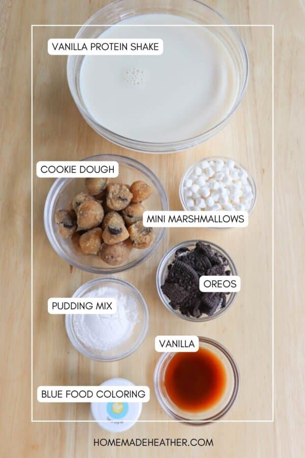 Ingredients for a vanilla protein shake are arranged on a wooden surface. Labeled bowls contain vanilla protein shake, mini marshmallows, cookie dough, Oreos, pudding mix, vanilla extract, and blue food coloring. homemadeheather.com is at the bottom.