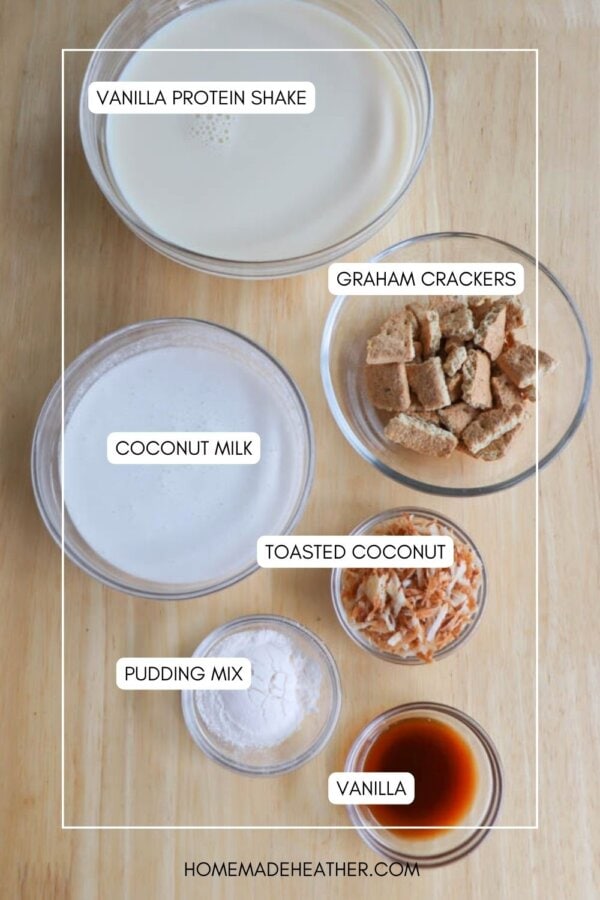 An overhead view of five labeled bowls on a wooden surface: vanilla protein shake, graham crackers, coconut milk, toasted coconut, pudding mix, and vanilla extract. Text at the bottom reads homemadeheather.com.