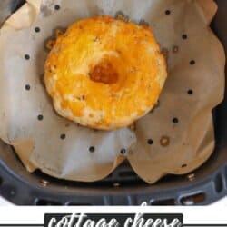 A cottage cheese bagel with melted cheese on top sits on parchment paper inside an air fryer basket. The image also includes text that reads cottage cheese air fryer bagels and homemadeheather.com.