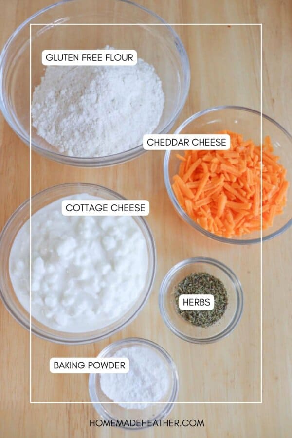 Top-down view of labeled ingredients in bowls on a wooden surface: gluten-free flour, cheddar cheese, cottage cheese, herbs, and baking powder.