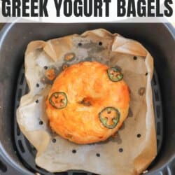 A jalapeรฑo cheddar bagel sits on parchment paper in a black air fryer basket. The bagel is topped with melted cheese and jalapeรฑo slices. Text above reads jalapeรฑo cheddar Greek yogurt bagels.