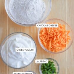 Top-down view of five bowls on a wooden surface. They contain gluten-free flour, cheddar cheese, Greek yogurt, jalapeรฑo, and baking powder. Each ingredient is labeled.