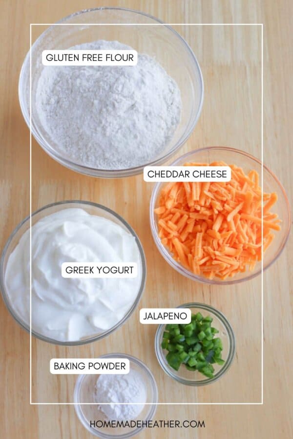 Top-down view of five bowls on a wooden surface. They contain gluten-free flour, cheddar cheese, Greek yogurt, jalapeรฑo, and baking powder. Each ingredient is labeled.