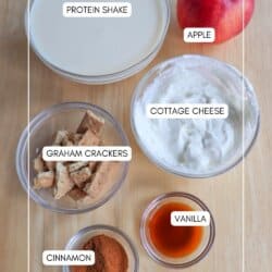 A wooden table with labeled ingredients: a protein shake, apple, cottage cheese, graham crackers, cinnamon, and vanilla. Each item is in a separate bowl or container, neatly arranged and labeled on the surface.