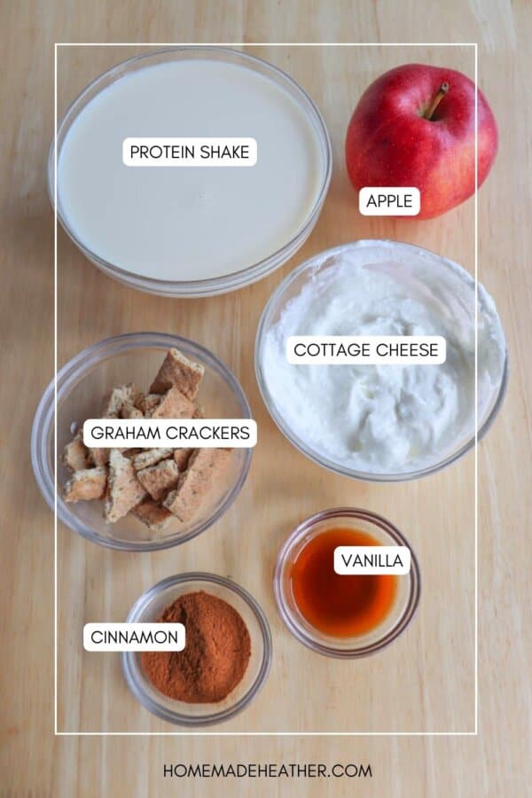 A wooden table with labeled ingredients: a protein shake, apple, cottage cheese, graham crackers, cinnamon, and vanilla. Each item is in a separate bowl or container, neatly arranged and labeled on the surface.