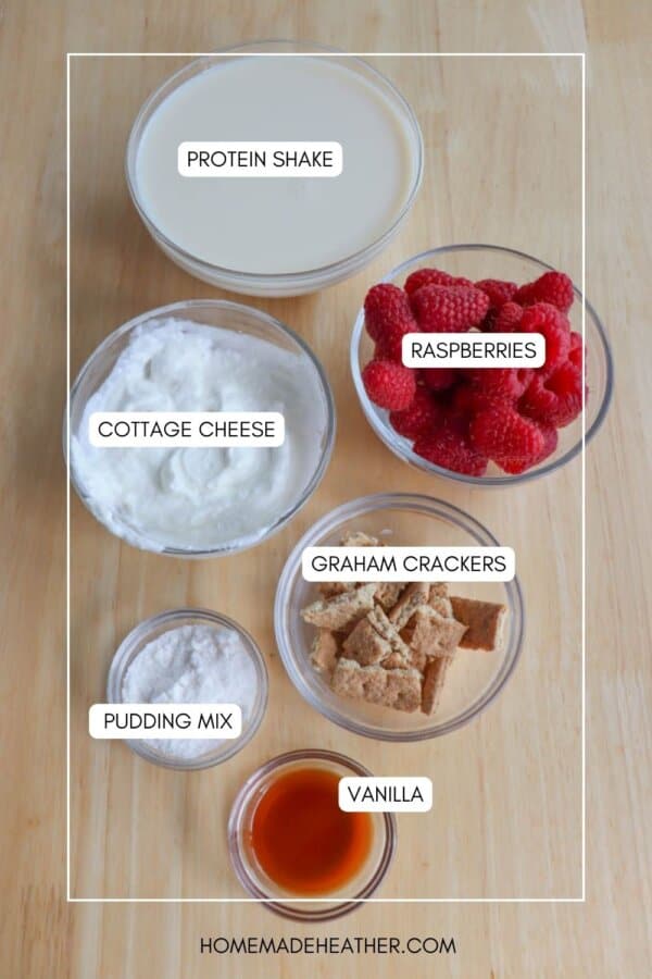 Top-down view of various ingredients in bowls on a wooden surface, labeled as protein shake, raspberries, cottage cheese, graham crackers, pudding mix, and vanilla.
