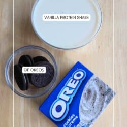 A glass of vanilla protein shake sits next to a small bowl containing gluten-free Oreos and an open box of Oreo pudding mix. They are arranged on a light wooden surface.