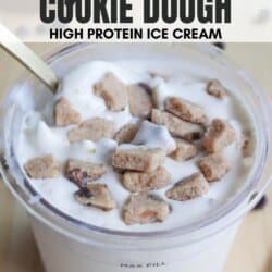 A tub of ninja creami cookie dough high protein ice cream is shown. It is filled close to the max fill line and topped with cookie dough chunks. A spoon is placed inside. The bottom displays the website homemadeheather.com.