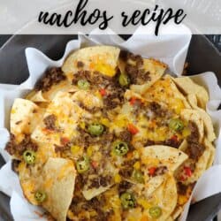 A close-up of nachos in a Dutch oven lined with parchment paper. The nachos are topped with melted cheese, ground beef, jalapeรฑos, and diced red peppers. A plaid cloth is partially visible in the background. Text reads Dutch Oven Nachos Recipe.