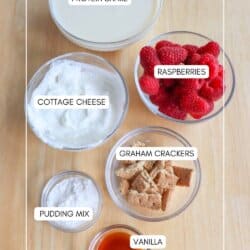 Ingredients for a recipe on a wooden surface: a bowl of protein shake, a bowl of cottage cheese, fresh raspberries reminiscent of blueberry cheesecake ice cream, graham crackers, pudding mix, and vanilla extract, each labeled accordingly.