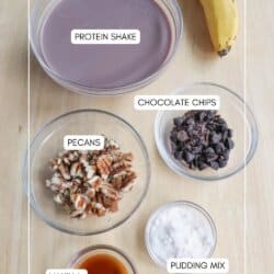 A flat lay of ingredients for a recipe, including a bowl of chocolate protein shake, a banana, and bowls of pecans, chocolate chips, vanilla extract, and pudding mix. Each ingredient is labeled.
