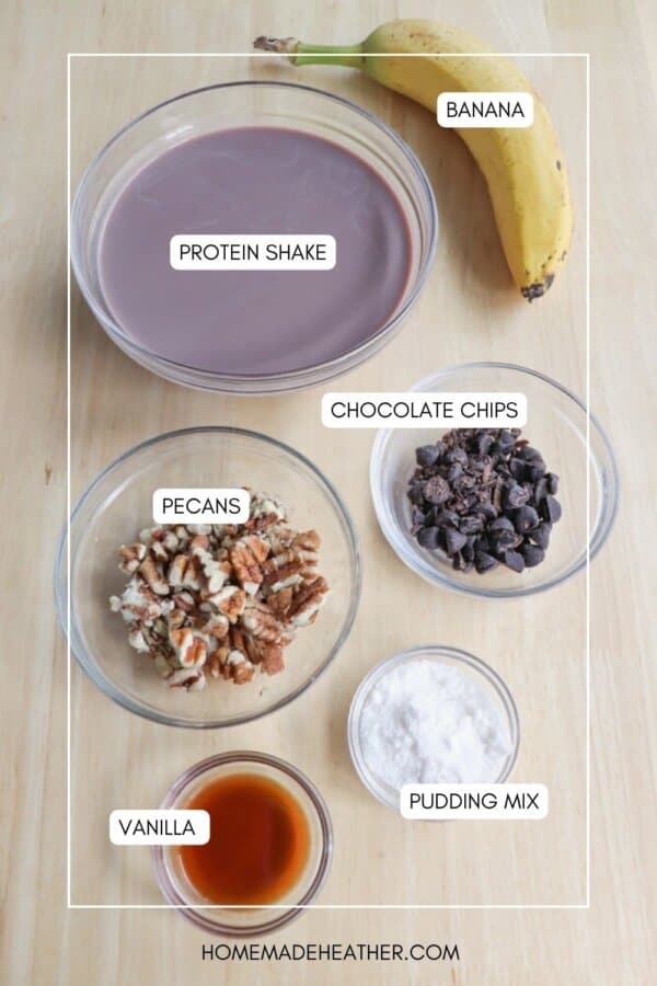 A flat lay of ingredients for a recipe, including a bowl of chocolate protein shake, a banana, and bowls of pecans, chocolate chips, vanilla extract, and pudding mix. Each ingredient is labeled.