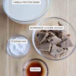 A top view of four labeled glass bowls on a wooden surface. They contain vanilla protein shake, homemade cookie dough, pudding mix, and vanilla. Labels describe each ingredient.