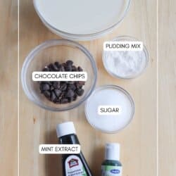 A top-view image of baking ingredients on a wooden surface, labeled: a bowl of vanilla protein shake, small bowls of pudding mix, chocolate chips, and sugar, alongside bottles of mint extract. Text at the bottom reads homemadeheather.com.