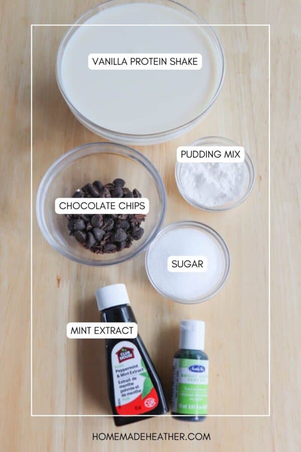A top-view image of baking ingredients on a wooden surface, labeled: a bowl of vanilla protein shake, small bowls of pudding mix, chocolate chips, and sugar, alongside bottles of mint extract. Text at the bottom reads homemadeheather.com.