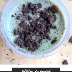 Top view of a bowl of mint ice cream topped with crumbled Oreo cookies. The text below reads ninja creami HIGH PROTEIN OREO MINT ICE CREAM homemadeheather.com. Oreo pieces are scattered on the wooden surface.