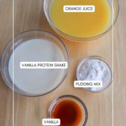 Bowls on a wooden surface hold the secret to a refreshing copycat Orange Julius recipe, with ingredients labeled: orange juice, vanilla protein shake, pudding mix, and vanilla extract.