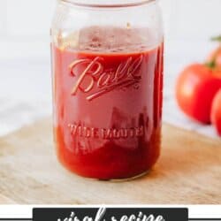 A glass jar filled with homemade ketchup sits on a wooden surface. The jar is labeled Ball and Wide Mouth. Two tomatoes are in the background. Text at the bottom reads: Viral Recipe - Homemade Ketchup - HomemadeHeather.com.