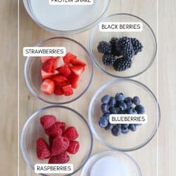 Top view of a wooden surface displaying bowls with labeled ingredients for a protein shake: sliced strawberries, blackberries, blueberries, raspberries, sugar, and a bowl of liquid labeled as protein shake.