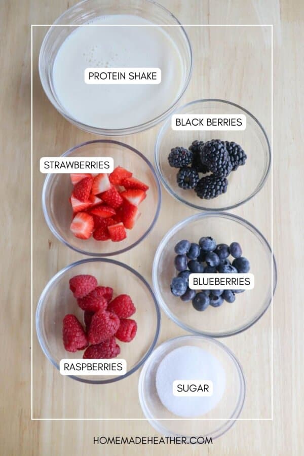 Top view of a wooden surface displaying bowls with labeled ingredients for a protein shake: sliced strawberries, blackberries, blueberries, raspberries, sugar, and a bowl of liquid labeled as protein shake.