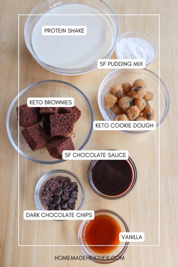 A wooden table displays various labeled ingredients in bowls: protein shake, SF pudding mix, keto brownies, keto cookie dough, SF chocolate sauce, dark chocolate chips, and vanilla. Text says HOMEMADEHEATHER.COM.