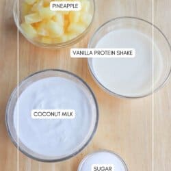 Top-down view of four bowls on a wooden surface containing pineapple chunks, vanilla protein shake, coconut milk, and sugar, each labeled with text indicating their contents.