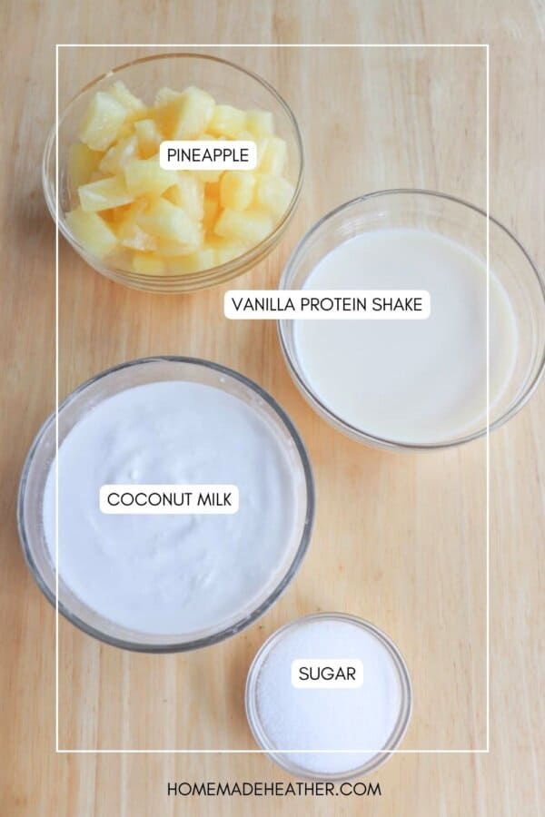 Top-down view of four bowls on a wooden surface containing pineapple chunks, vanilla protein shake, coconut milk, and sugar, each labeled with text indicating their contents.