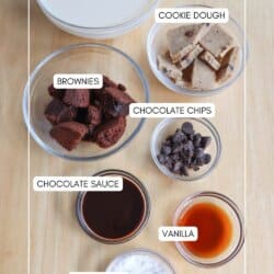 A wooden table holds ingredients for a protein shake recipe. Includes labeled bowls of cookie dough, chocolate chips, brownies, chocolate sauce, vanilla, pudding mix, and a glass of protein shake.