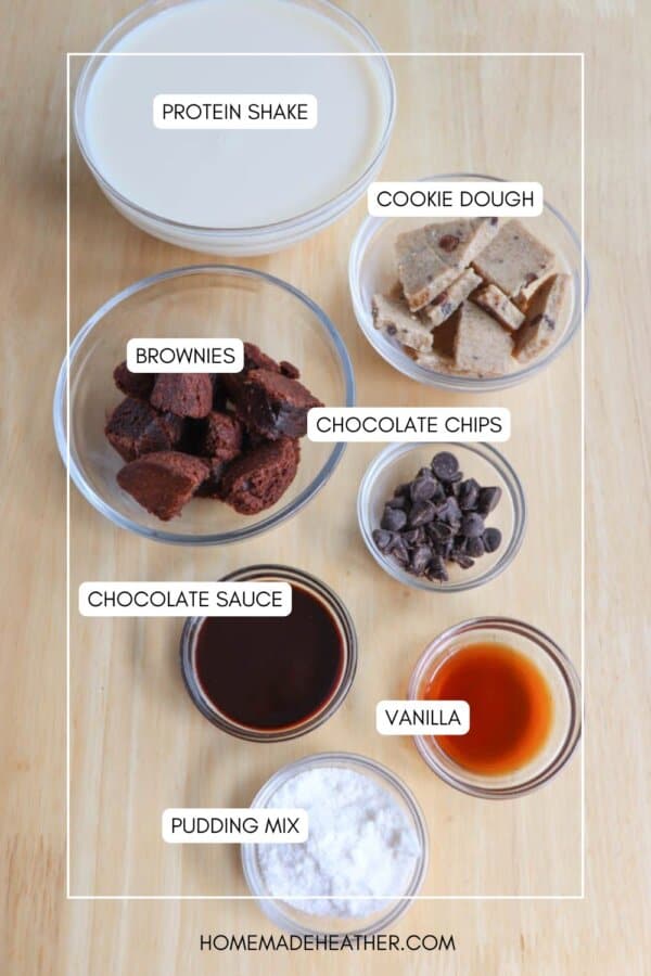 A wooden table holds ingredients for a protein shake recipe. Includes labeled bowls of cookie dough, chocolate chips, brownies, chocolate sauce, vanilla, pudding mix, and a glass of protein shake.