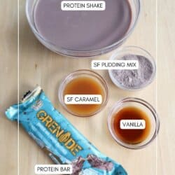A bowl of protein shake, small labeled bowls of SF pudding mix, SF caramel, and vanilla, and a packaged protein bar are arranged on a wooden surface.