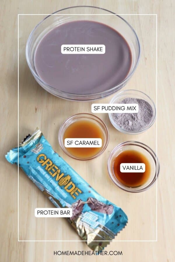 A bowl of protein shake, small labeled bowls of SF pudding mix, SF caramel, and vanilla, and a packaged protein bar are arranged on a wooden surface.