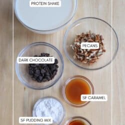 Top-down view of ingredients on a wooden surface, including labeled items: protein shake, pecans, dark chocolate, sugar-free caramel, sugar-free pudding mix, and vanilla. Containers are arranged with text labels and a website watermark at the bottom.