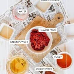 A flat lay of chili sauce ingredients on a wooden board and newspaper. Tomato paste in a bowl, surrounded by labeled bowls: garlic powder, onion powder, chili powder, water, honey, and apple cider vinegar.