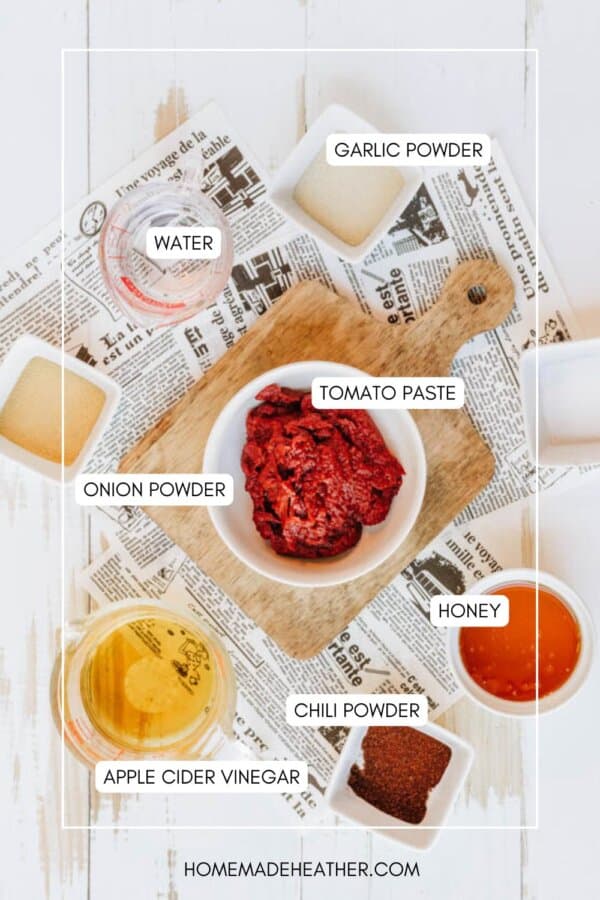 A flat lay of chili sauce ingredients on a wooden board and newspaper. Tomato paste in a bowl, surrounded by labeled bowls: garlic powder, onion powder, chili powder, water, honey, and apple cider vinegar.