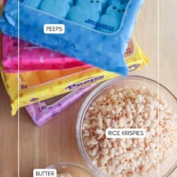 Packages of Peeps in yellow, pink, and blue are stacked beside a bowl of Rice Krispies and a small bowl of butter on a wooden surface.