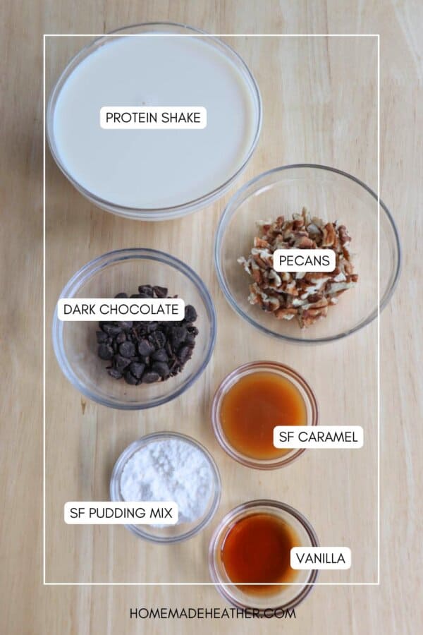 Top-down view of ingredients on a wooden surface, including labeled items: protein shake, pecans, dark chocolate, sugar-free caramel, sugar-free pudding mix, and vanilla. Containers are arranged with text labels and a website watermark at the bottom.