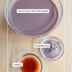 A bowl of chocolate protein shake, a small bowl of pudding mix, and a smaller bowl of vanilla extract are placed on a wooden surface. Labels identify each ingredient.