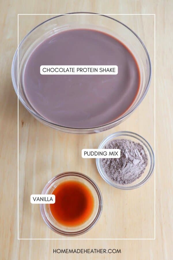A bowl of chocolate protein shake, a small bowl of pudding mix, and a smaller bowl of vanilla extract are placed on a wooden surface. Labels identify each ingredient.