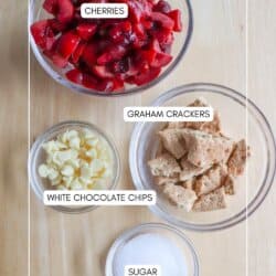 Four glass bowls on a wooden surface contain chopped cherries, broken graham crackers, white chocolate chips, and sugar. Each ingredient is labeled.