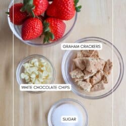 A top view of ingredients for a recipe on a wooden surface. A bowl of fresh strawberries, a bowl of graham cracker pieces, white chocolate chips in a small bowl, and sugar in another small bowl.