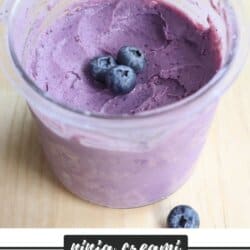 A clear glass bowl filled with creamy purple blueberry frozen yogurt, topped with three whole blueberries. The background shows a wooden surface. At the bottom, theres text: ninja creami BLUEBERRY FROYO homemadeheather.com.