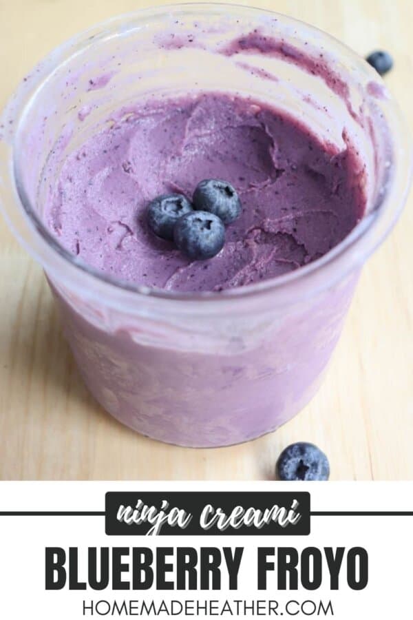 A clear glass bowl filled with creamy purple blueberry frozen yogurt, topped with three whole blueberries. The background shows a wooden surface. At the bottom, theres text: ninja creami BLUEBERRY FROYO homemadeheather.com.