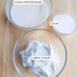 Three glass bowls on a wooden surface: one with vanilla protein shake, another with sugar, and the last with Greek yogurt. Each bowl is labeled accordingly.