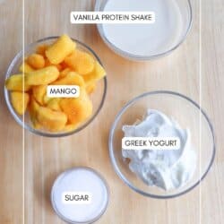 Four bowls on a wooden table: one with vanilla protein shake, one with diced mango, one with Greek yogurt, and a small one with sugar. Each bowl is labeled accordingly.