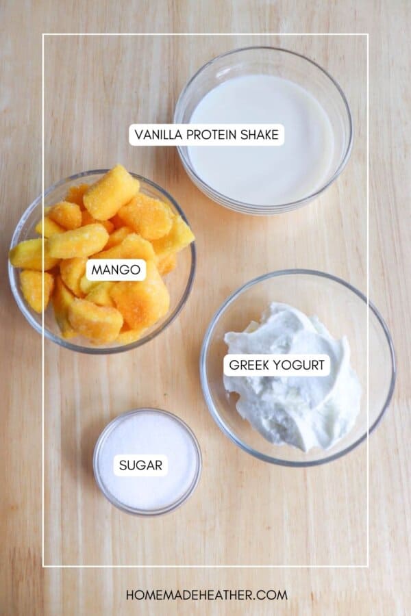Four bowls on a wooden table: one with vanilla protein shake, one with diced mango, one with Greek yogurt, and a small one with sugar. Each bowl is labeled accordingly.