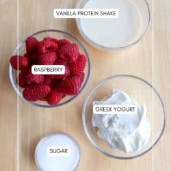 Top-down view of four bowls on a wooden surface labeled with ingredients for a protein shake: one with milk labeled Vanilla Protein Shake, one with raspberries, one with Greek yogurt, and a small bowl of sugar.